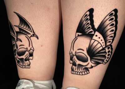 American traditional side by side skulls with wings tattoo in black gray on front of shins